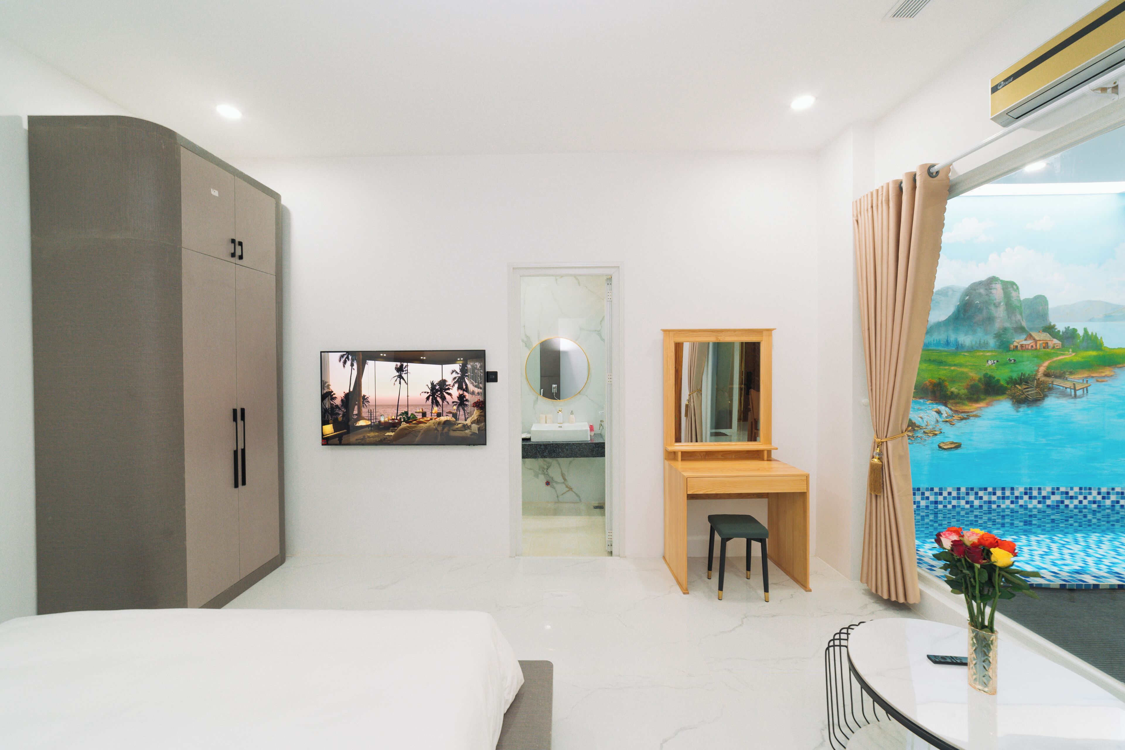 A modern hotel room with a bed, a desk, a mirror, a bathroom with a sink, and a large window with a view of a landscape.
