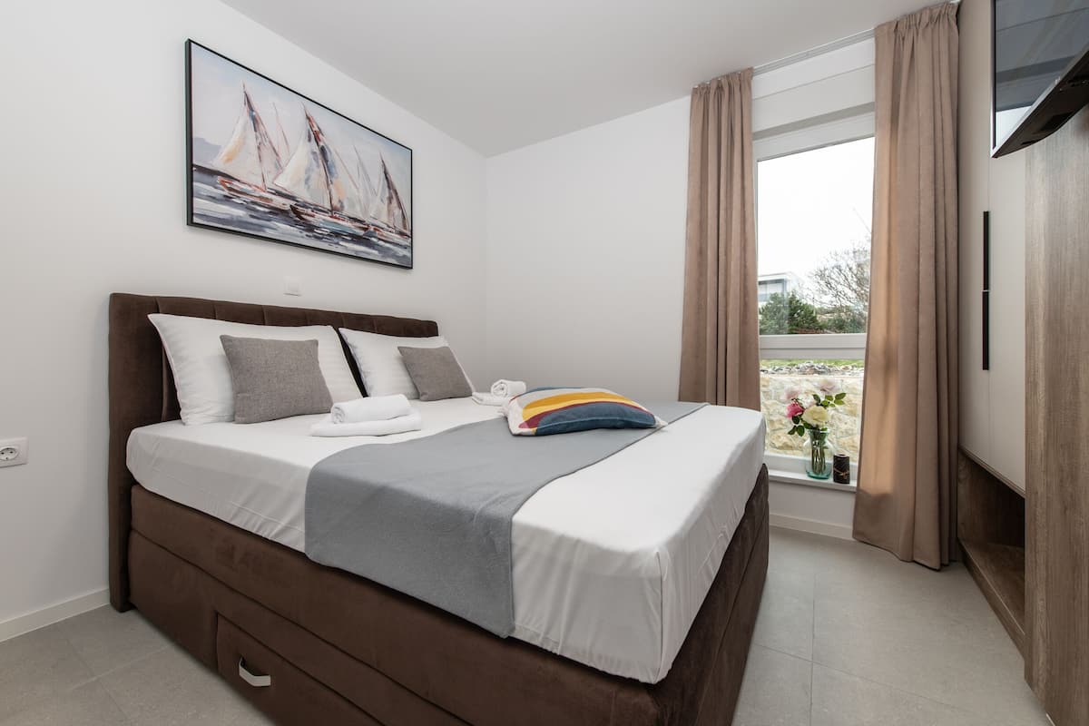 A neatly made bed with a gray and white duvet, a colorful pillow, and a white pillow. There is a painting of sailboats on the wall above the bed.
