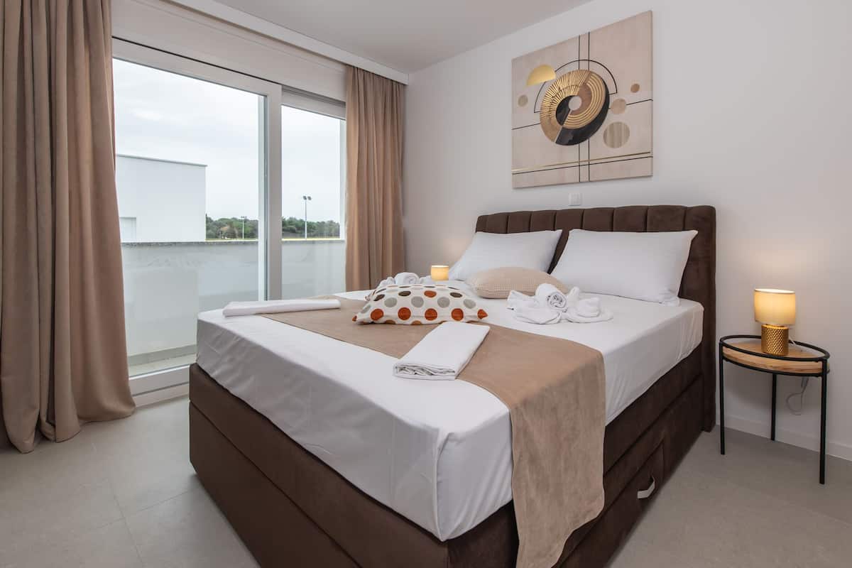 A neatly made bed with a brown headboard, a white pillow, and a polka-dotted pillow. There is a small bedside table with a lamp and a towel.