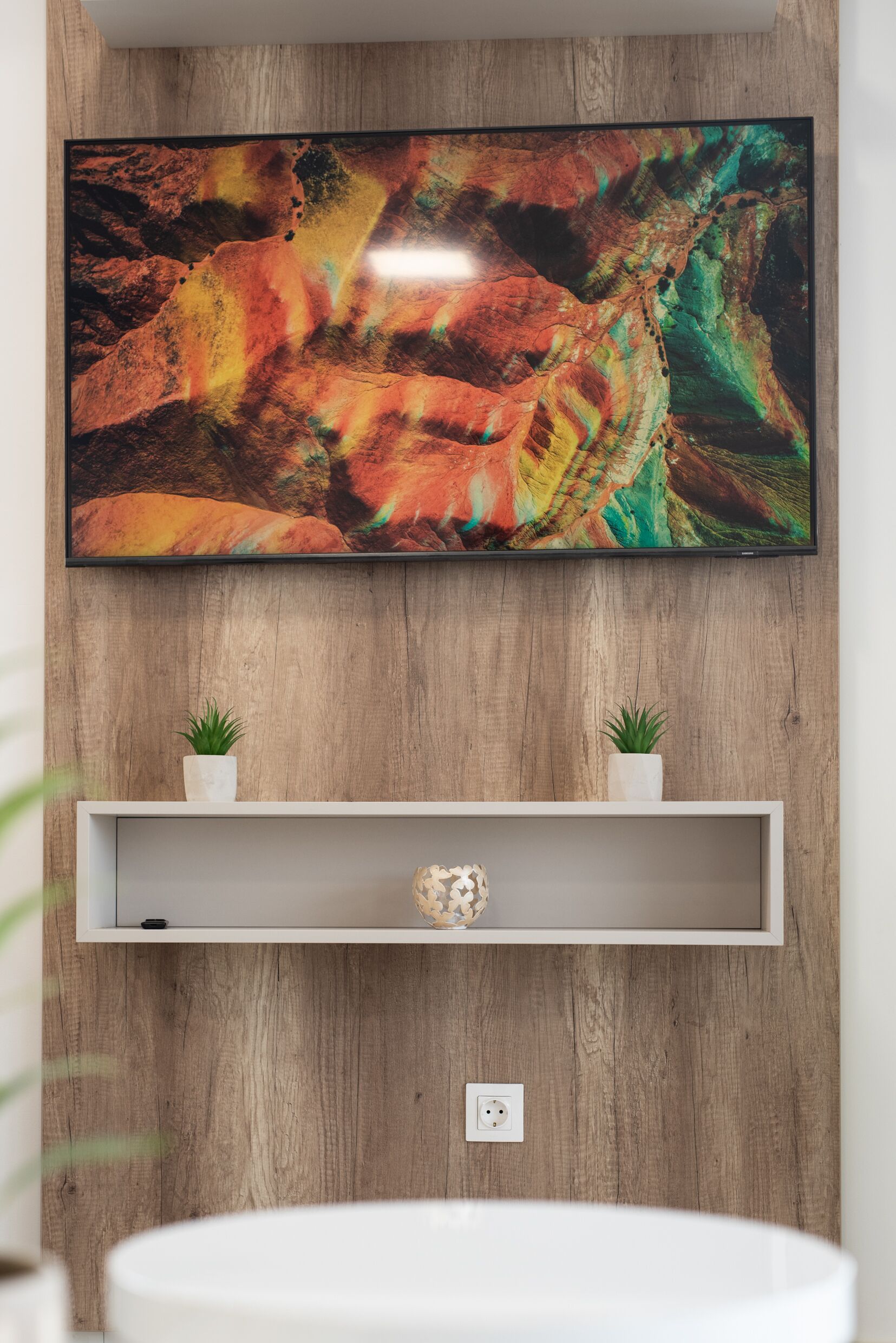 Una TV a schermo piatto montata su un parete di legno sopra un ripiano bianco con due piante in vaso.