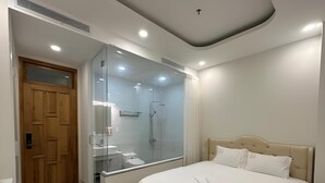 A modern bedroom with a glass-enclosed shower, a wooden door, a bed with white bedding, and a small bedside table. - Hala Hotel - by Bay Luxury (Ho Chi Minh City)