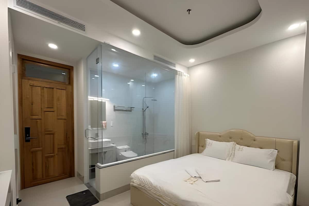A modern bedroom with a glass-enclosed shower, a wooden door, a bed with white bedding, and a small bedside table.