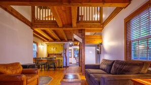 A cozy living room with wooden floors, a leather sofa, a dark armchair, and a wooden coffee table. A kitchen with wooden cabinets and a dining area with chairs are visible in the background. - Private Sauna · Oceanfront Cabin w/ Views! Steps to the Beach! ⎮Hidden Pearl (Ucluelet)