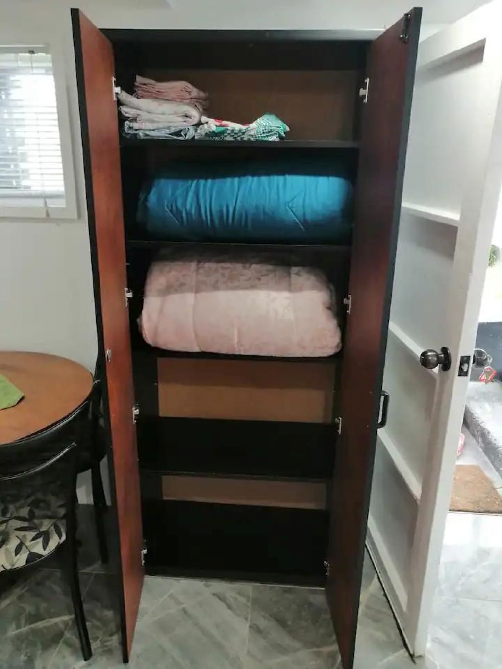 Een inloopkast met verschillende planken, een tafeltje met een stoel, en een deur naar een andere kamer.