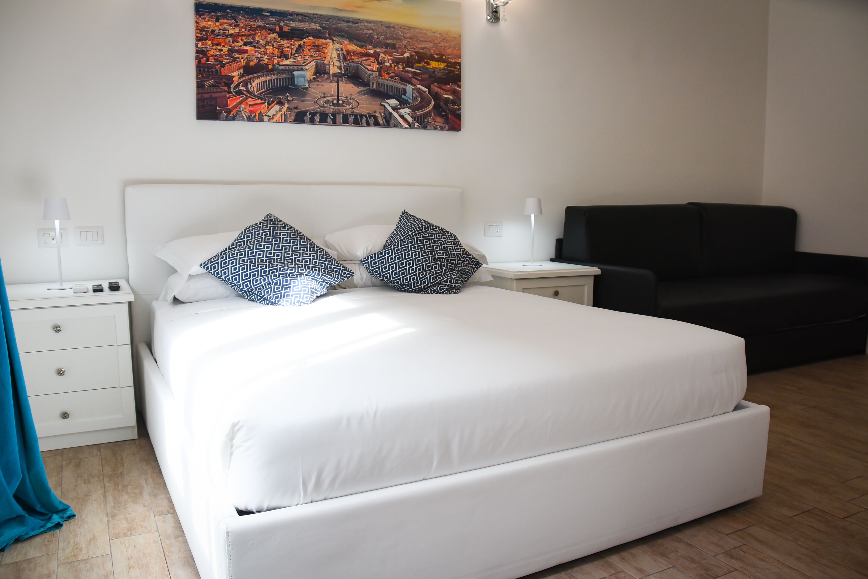 A neatly arranged bedroom with a large bed, a black sofa, and a cityscape painting on the wall.