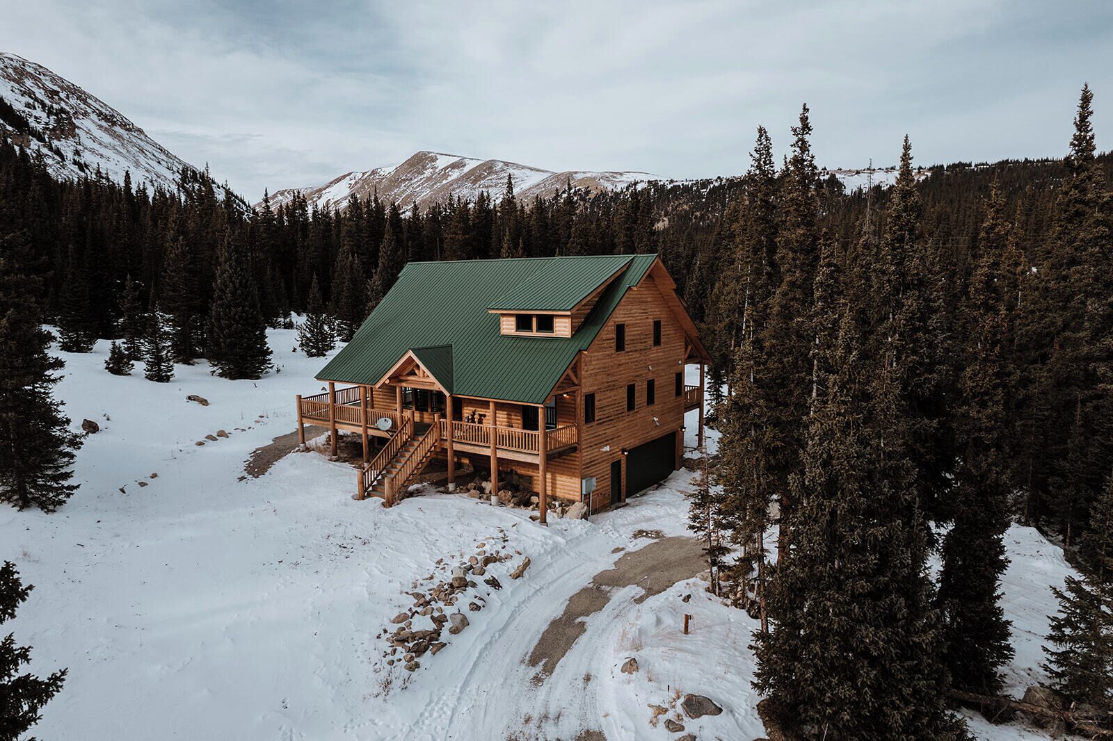 A log cabin with a green roof, surrounded by snow-covered ground and dense forest, with mountains in the background.