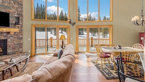 A spacious living room with a stone fireplace, large windows, a dining area, and a view of a snowy landscape. - Log Cabin on 10 Acres with Loft and Soaker Tub. (Fairplay)