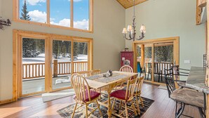 A spacious dining area with a wooden table and chairs, large windows with a view of a snowy deck, and a chandelier. - Log Cabin on 10 Acres with Loft and Soaker Tub. (Fairplay)