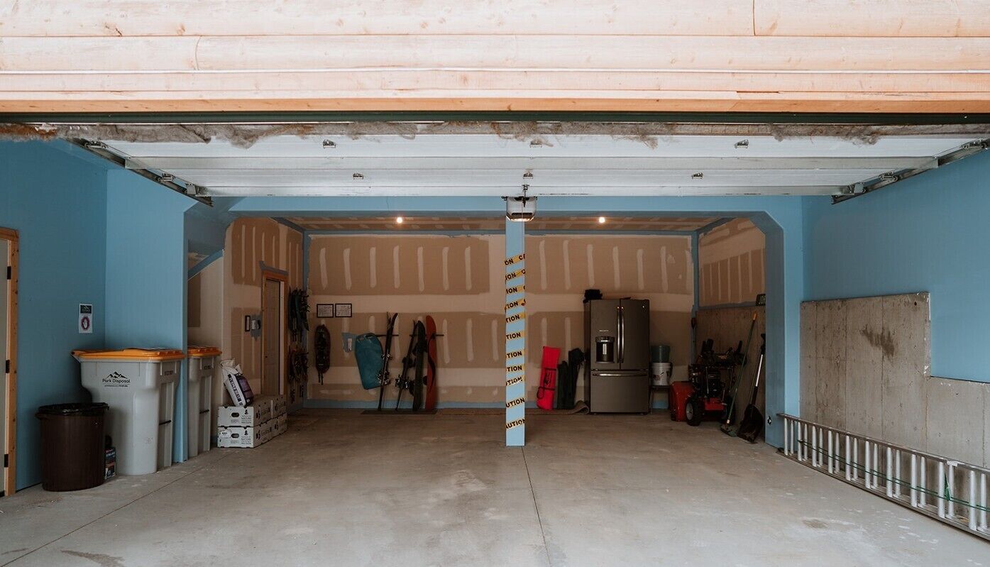 A garage with a concrete floor, a wooden door, and a blue wall. There are two trash bins, a refrigerator, and a red lawnmower visible.