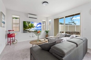 A modern living room with a grey sectional sofa, a flat-screen TV, a red cart, and a large window with a view of the outdoors. - @ Marbella Lane - Stunning Gem w/ MT & Ocean Views (Waianae)