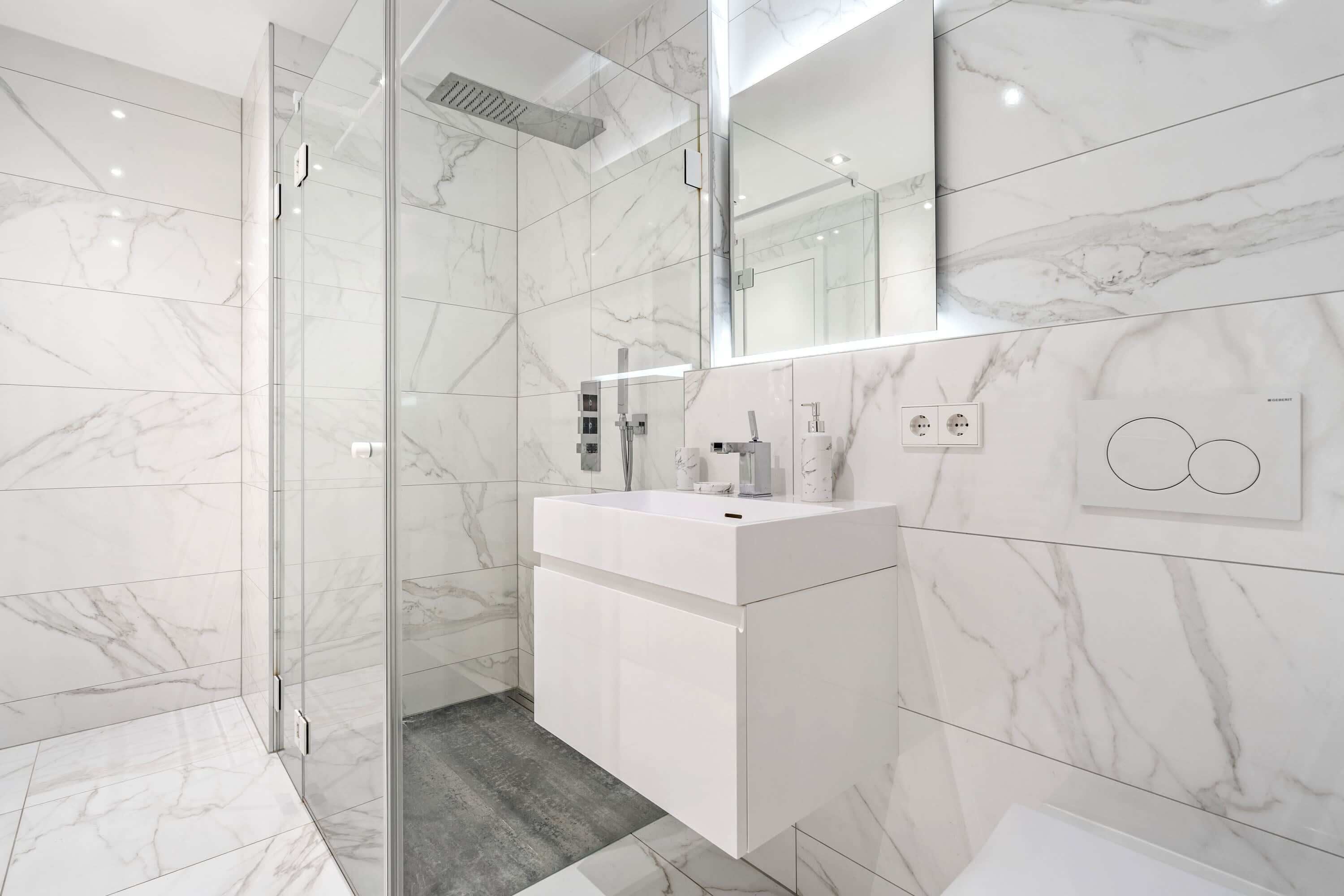 Un bany modern amb un box de dutxa de vidre, un lavabo blanc amb mirall i parets amb disseny de marbre.