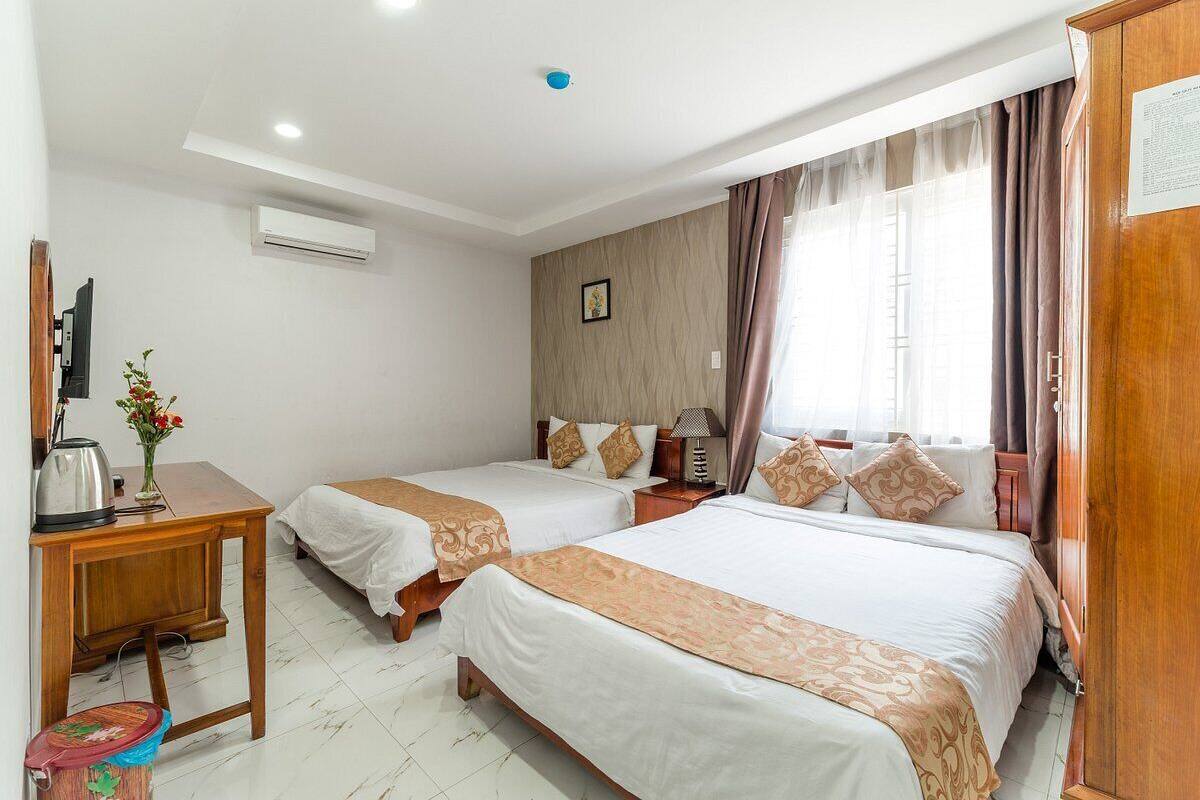 A hotel room with two beds, a wooden desk, a trash bin, an air conditioning unit, and a window with curtains.
