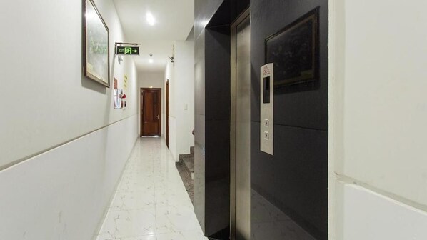 A hallway with a marble floor, a door with an elevator button, and an exit sign. - Rose Hotel  (Da Nang)