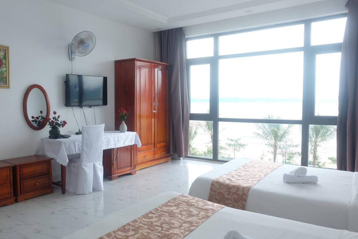 A hotel room with a large window, a TV, a wardrobe, a desk with a mirror, and two beds.