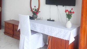 Ein Zimmer mit Schreibtisch aus Holz, einem Sessel, einem an der Wand befestigten Fernseher, einem kleinen Tisch mit Blumenvase und einem Spiegel.