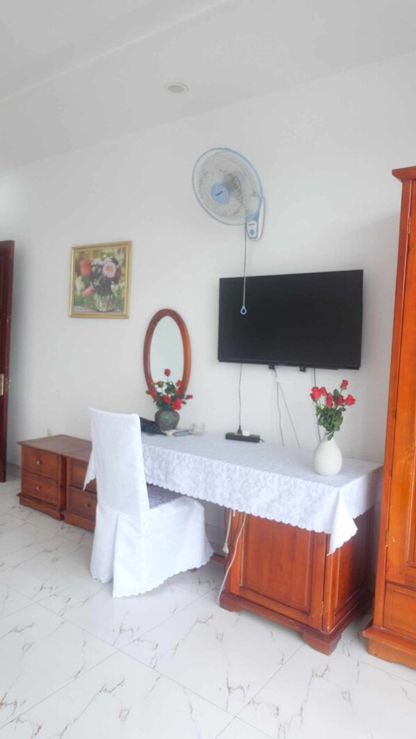Una habitación con un escritorio de madera, una silla, un televisor montado en la pared, una mesita con un florero y un espejo.