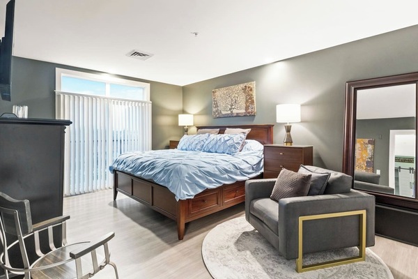 This bedroom offers a cozy atmosphere with its warm gray walls and complementing dark wood furniture, including a large bed with blue bedding. A modern armchair with a gold frame, a circular rug, and artistic wall hangings complete the inviting and s