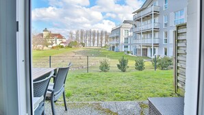 Une vue depuis un balcon donnant sur un espace verdoyant, avec une clôture, un immeuble doté de plusieurs balcons et, au loin, un bâtiment avec une tour.