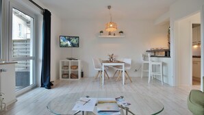 Un salon moderne avec une table basse en verre, des meubles blancs, un téléviseur fixé au mur et un coin cuisine visible à travers une porte ouverte.