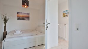 Une chambre à coucher avec un lit, une lampe fixée au mur, un tableau représentant un coucher de soleil et un radiateur.