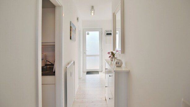 Un couloir étroit aux murs blancs et au plancher en bois clair. Sur la gauche, on trouve un coin cuisine avec un évier et des placards. Un miroir et un vase de fleurs sont placés sur le côté droit.