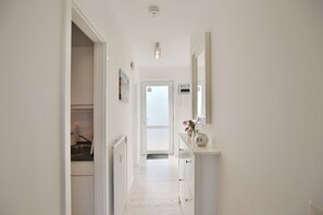 Ein schmaler Flur mit weißen Wänden und Holzdielen. Links befindet sich eine Küchenzeile mit Spüle und Schränken. Rechts stehen ein Spiegel und eine Vase mit Blumen.