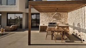A modern outdoor patio with a stone wall, wooden pergola, and a built-in grill station. - Mayamor Villas (Zakynthos)