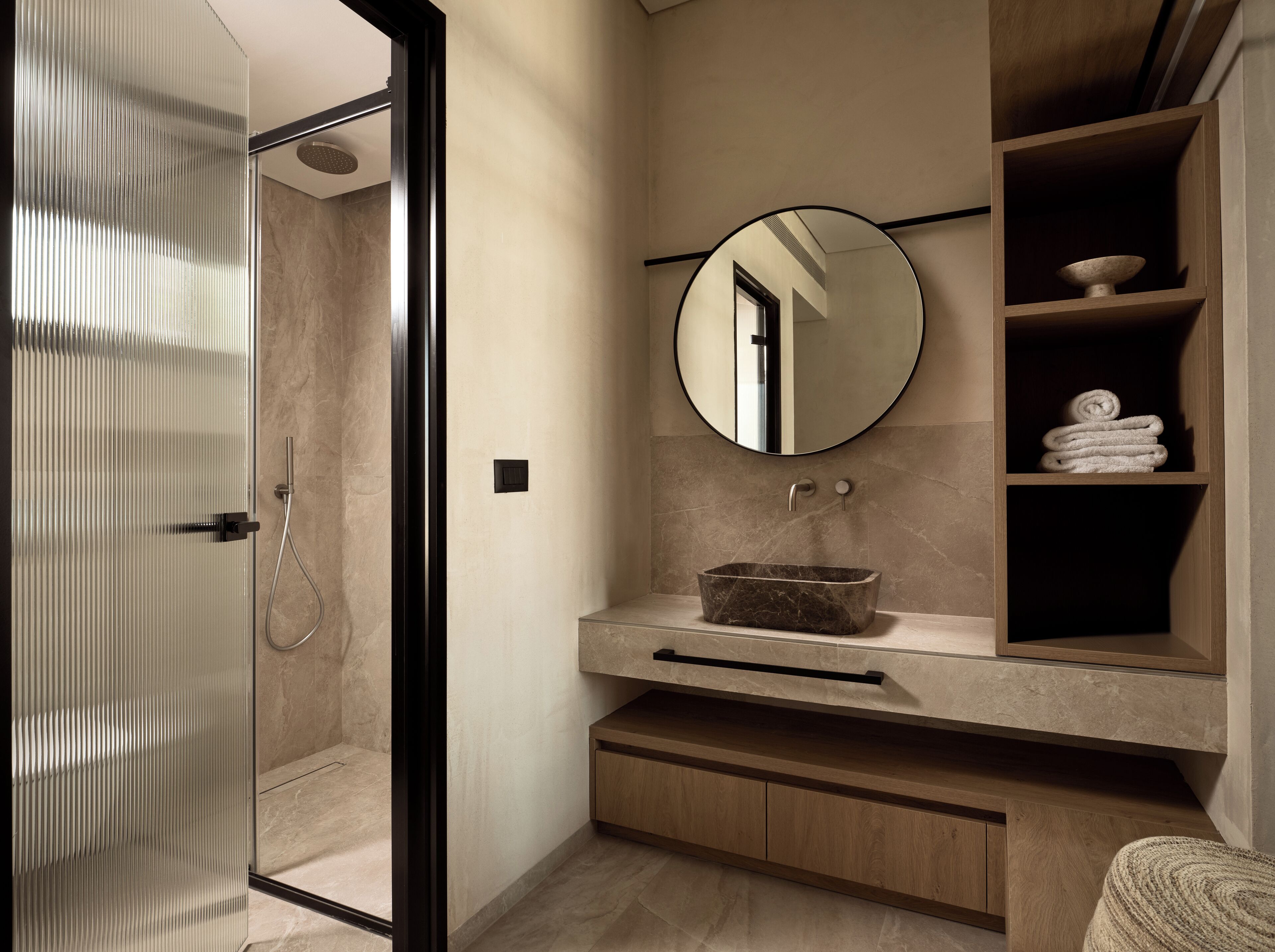A modern bathroom with a glass shower enclosure, a round mirror, a marble sink, and a wooden vanity with a towel rack.
