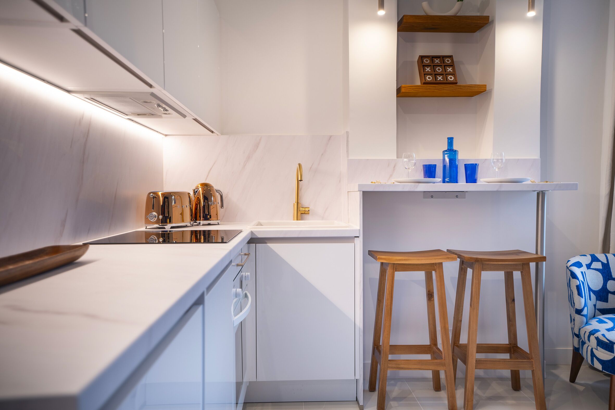 A modern kitchen with white cabinetry, a marble backsplash, and a wooden countertop. There are two wooden stools and a blue chair.