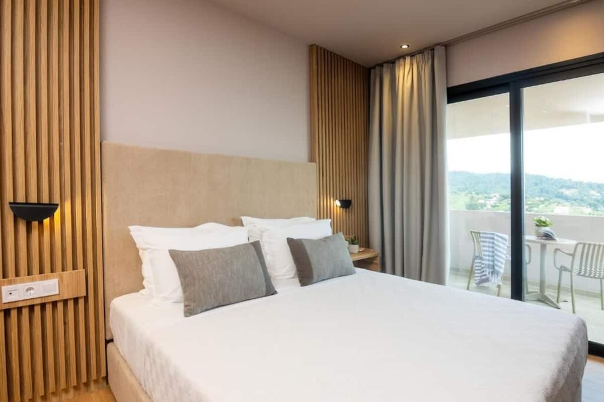 A modern hotel room with a large bed, a wooden headboard, a nightstand, and a balcony with outdoor furniture.