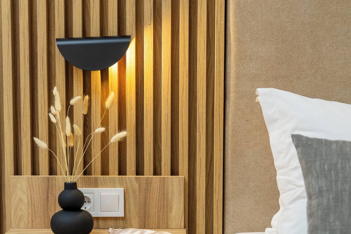 A modern bedroom with a wooden slat wall, a bedside table with a black lamp, a vase with dried pampas grass, and a bed with white and gray bedding.