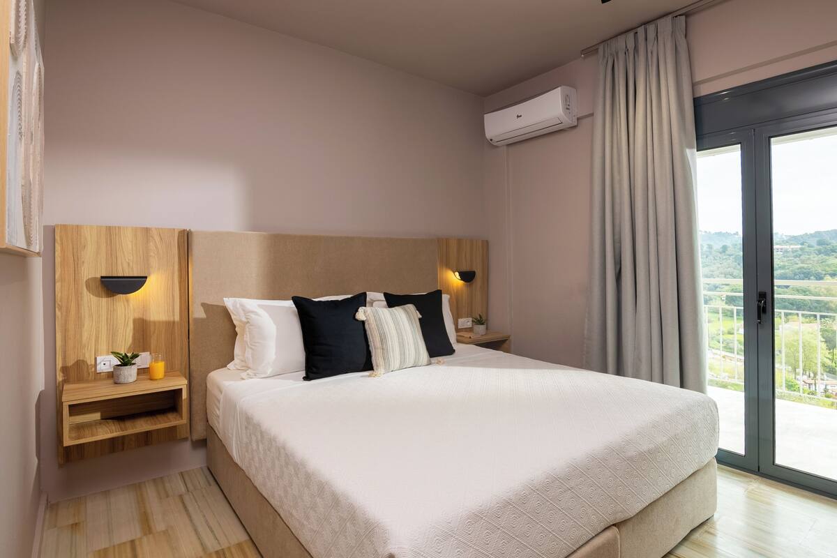 A hotel room with a large bed, wooden headboard, bedside tables with lamps, and a view of greenery through a sliding glass door.