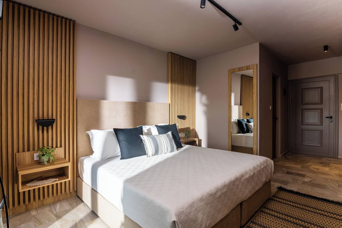 A modern bedroom with a large bed, wooden paneling, a bedside table with a plant, and a mirror above the bed.