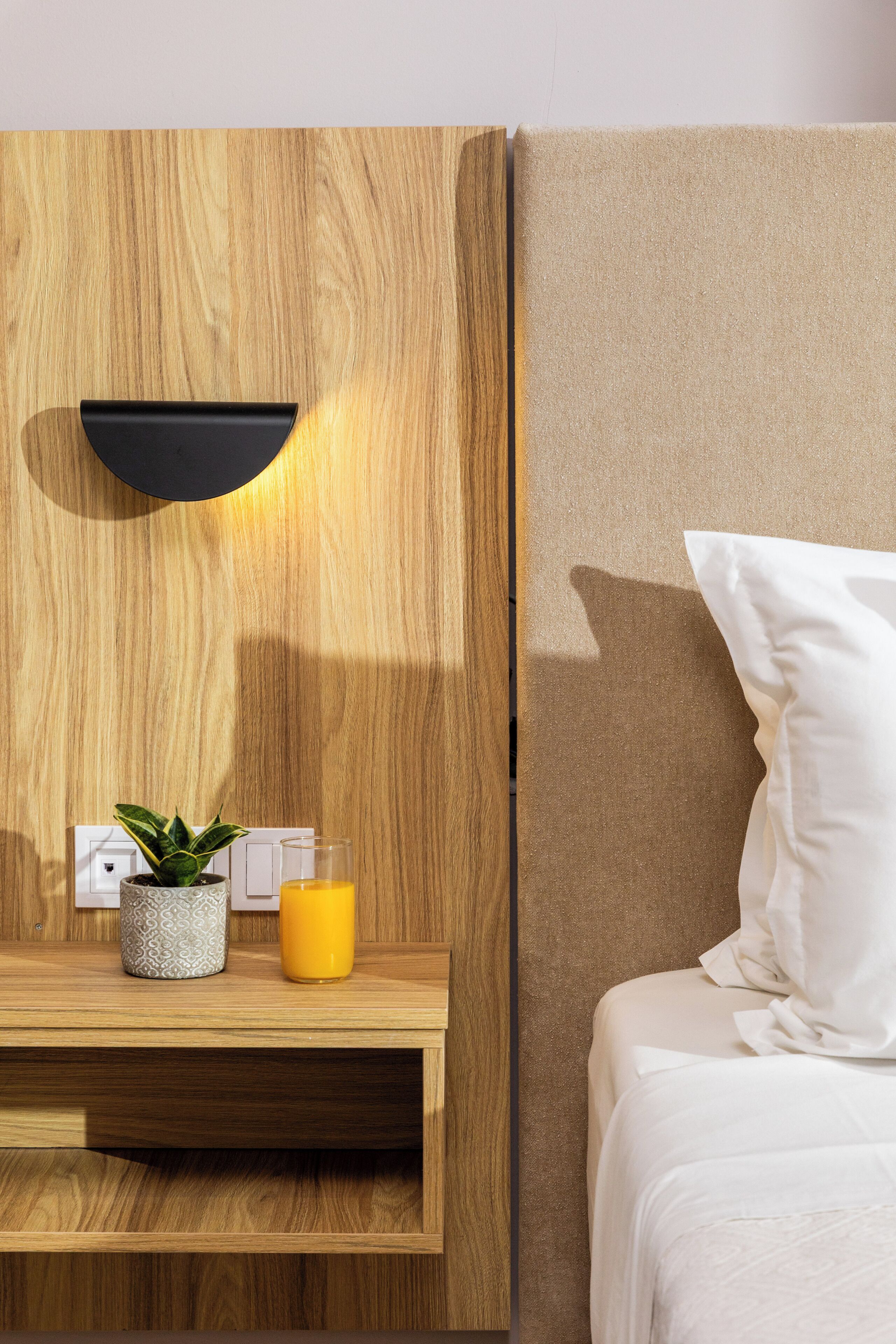 A modern bedroom with a wooden headboard, a bedside table with a plant and a glass of orange juice, and a white pillow on the bed.