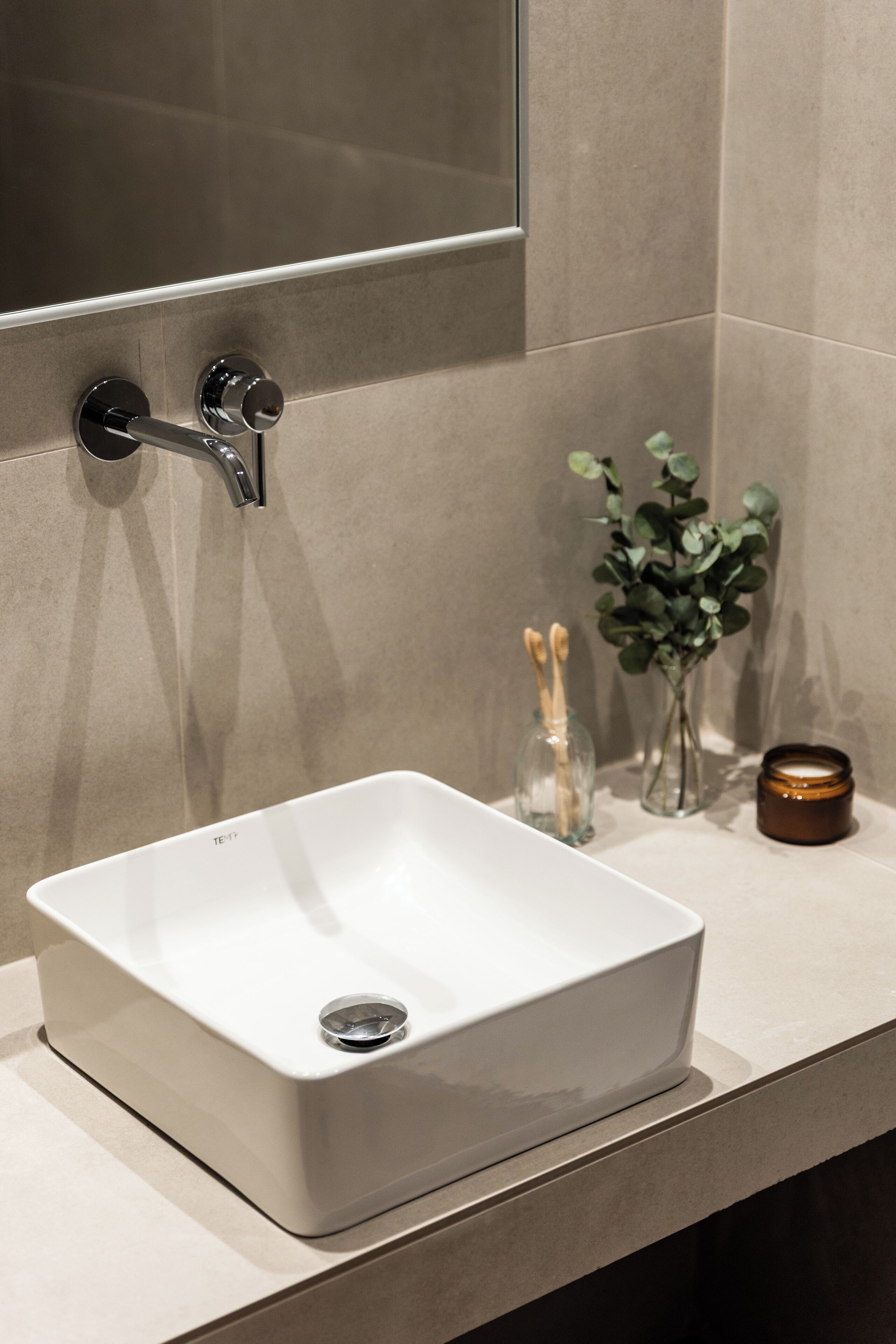 Une salle de bain avec un lavabo blanc, un miroir et un petit vase contenant des feuilles vertes.