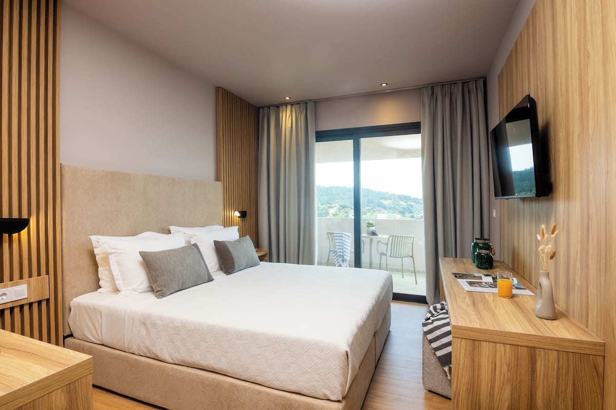 A modern hotel room with a large bed, a flat-screen TV, a wooden desk, and a balcony with a view.