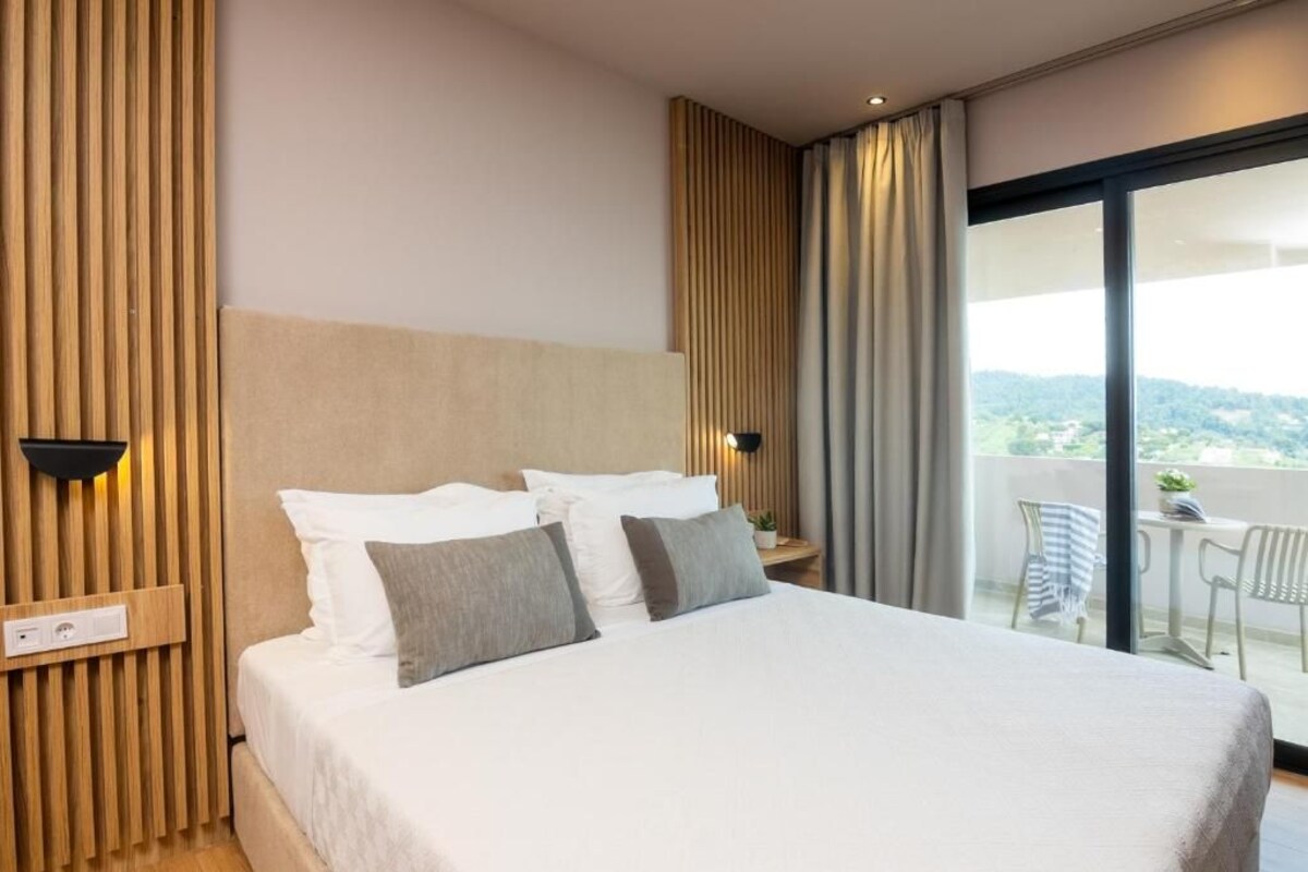 Une chambre d’hôtel moderne dotée d’un grand lit, d’une tête de lit en bois, d’une table de chevet et d’un balcon aménagé avec du mobilier d’extérieur.