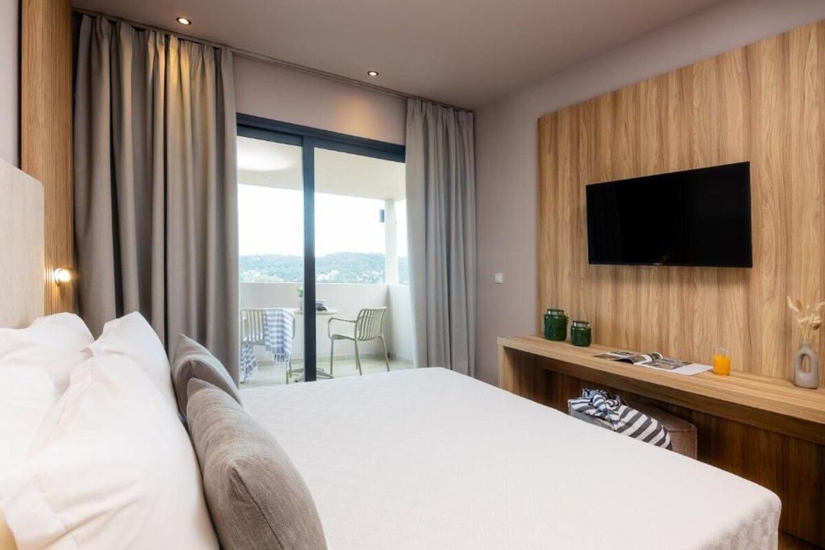 Une chambre d’hôtel avec un lit, un téléviseur fixé au mur en bois, un balcon avec une table et une chaise, et une vue sur l’extérieur.