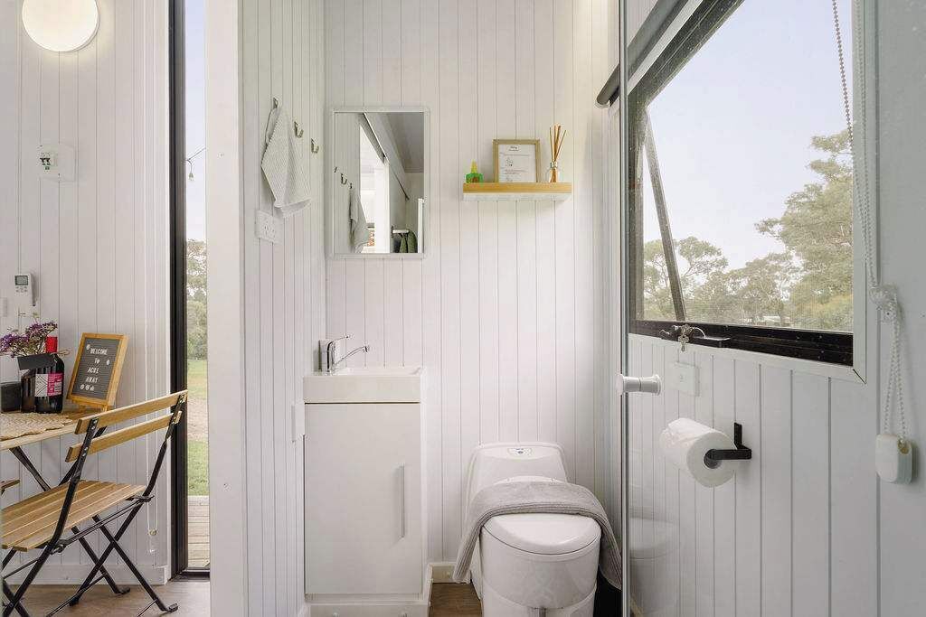 Une petite salle de bain comprenant des toilettes, un lavabo et un miroir. On y trouve une petite étagère avec des articles de toilette et un panneau d’accueil encadré.
