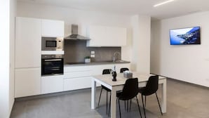Une cuisine moderne avec des meubles blancs, un four encastré et un four à micro-ondes. On y trouve une table à manger avec des chaises et un téléviseur à écran plat fixé au mur.
