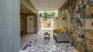 Un espacio al aire libre cubierto con pared de piedra, una puerta blanca, un sofá de mimbre con cojines y suelo de baldosas.