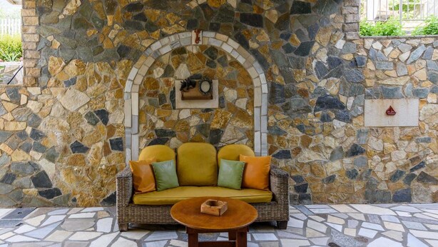 Un muro de piedra con un diseño de mosaico, un sofá de mimbre con cojines y una mesita.