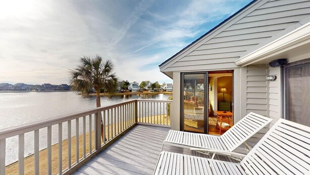 A wooden deck with two white lounge chairs, a glass door leading to an interior room, and a view of a body of water and distant buildings. - Views of Osprey Lake in Pelican Watch 4 Bedroom (Pawleys Island)