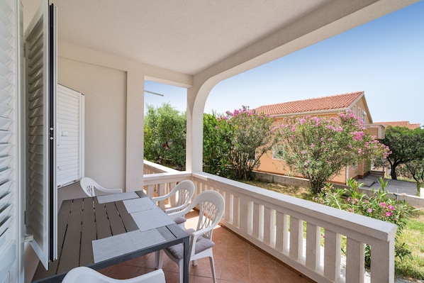 A balcony with a table and chairs, a white railing, and a view of a building with a red roof and pink flowers.