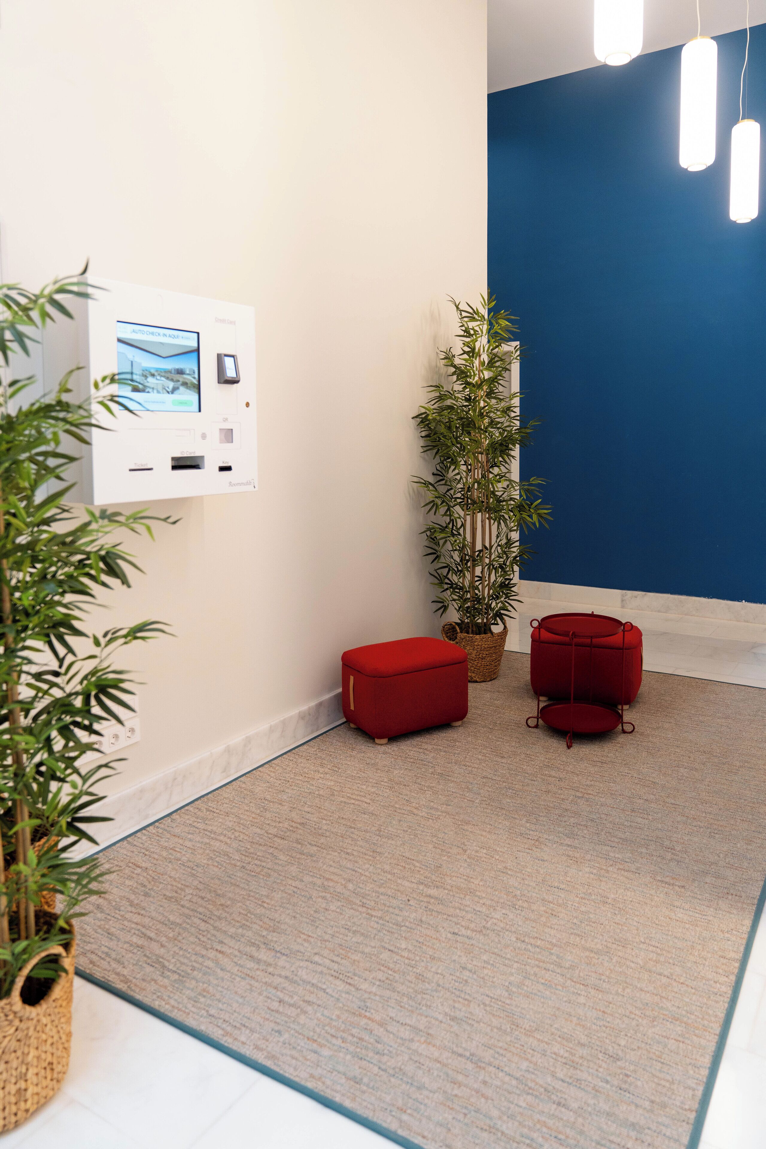 A room with a wall-mounted control panel, a red ottoman, a tall potted plant, and a red chair.