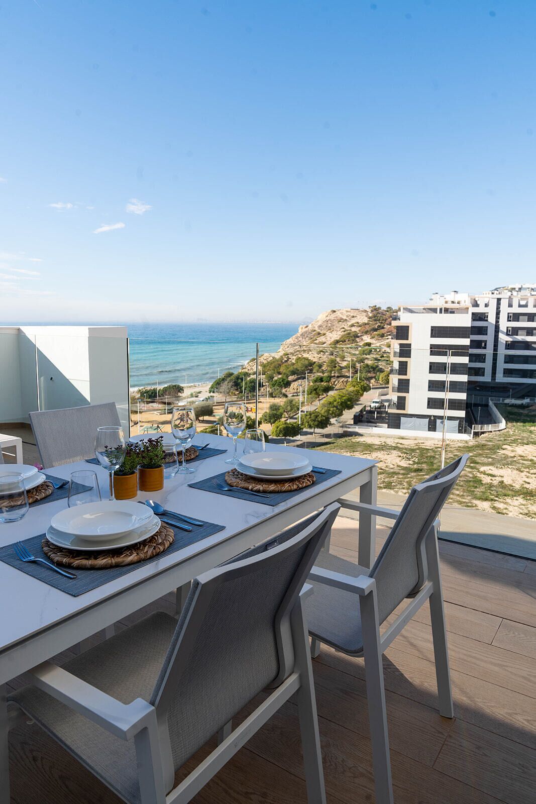 A dining table set with plates, glasses, and cutlery on a balcony overlooking a beach and buildings.