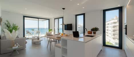 A modern kitchen with a central island, dining area with chairs, and large windows offering a view of the sea.