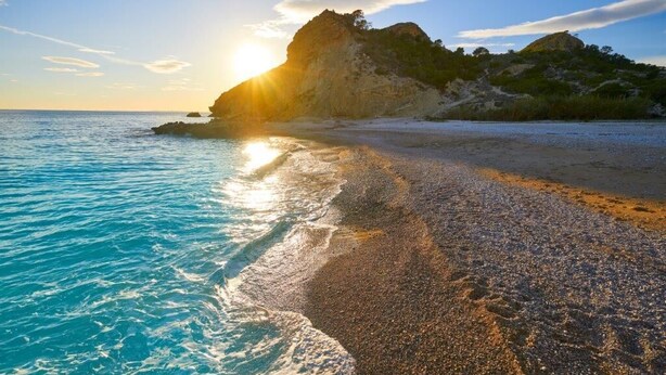 A beach with clear turquoise water, pebbly shore, and a rocky cliff with vegetation. - Horizonte Line (Villajoyosa)