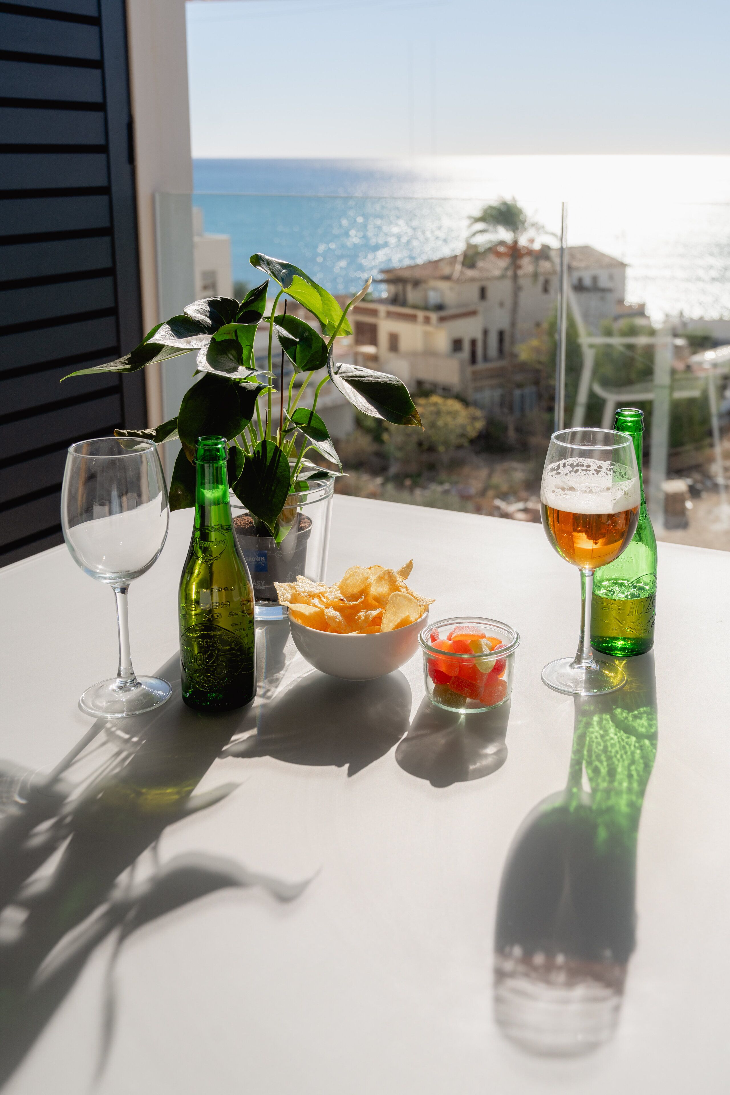 Une table avec une bouteille verte, un verre à vin, un bol de chips et un verre de bière, avec une vue sur la mer et des bâtiments en arrière-plan.