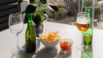 Une table avec une bouteille verte, un verre à vin, un bol de chips et un verre de bière, avec une vue sur la mer et des bâtiments en arrière-plan.